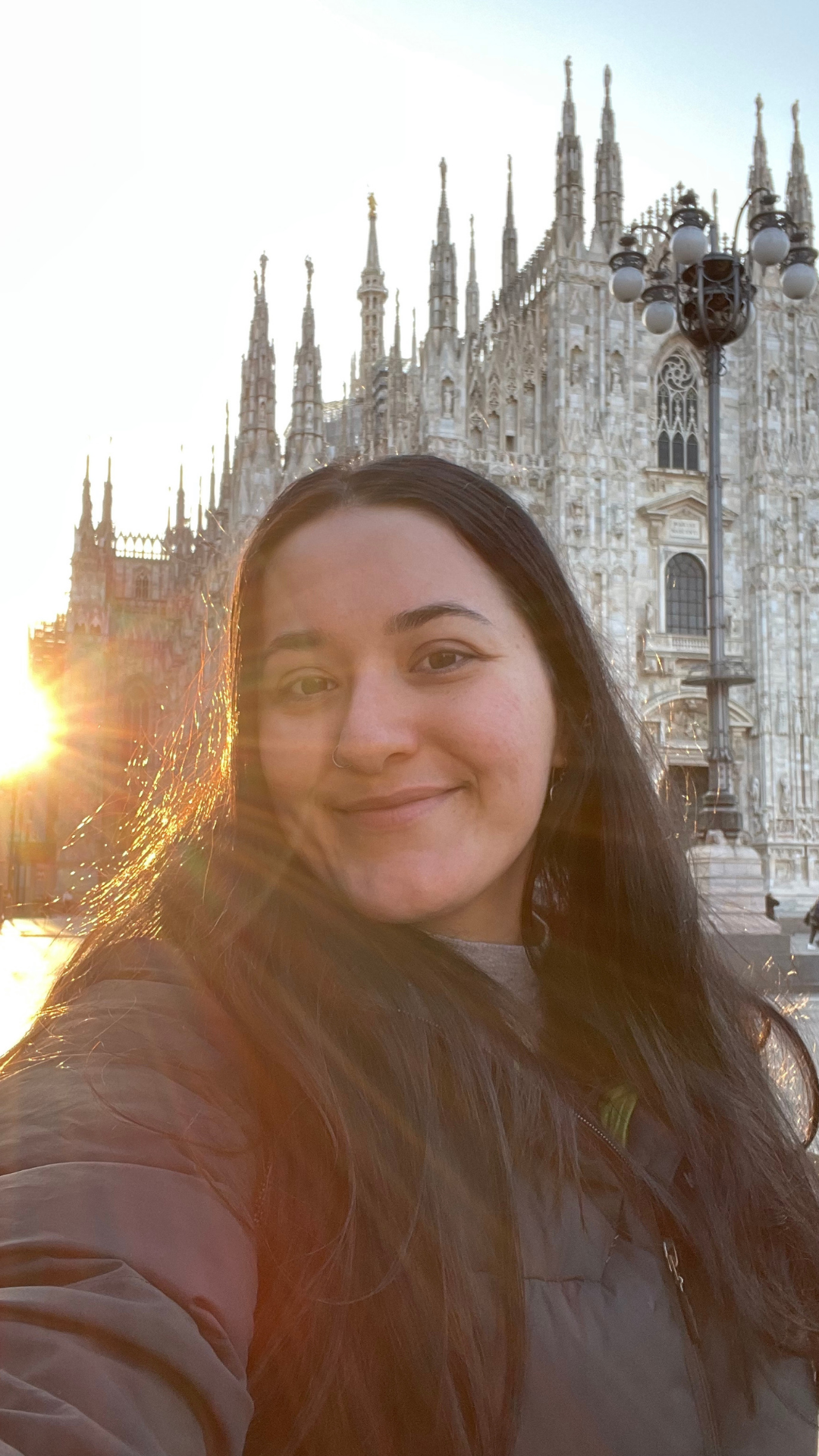 Silhouetted against the stunning sunrise, a figure stands in the foreground, their face gently illuminated by the warm, golden hues of the morning light. In the distance, the magnificent Duomo di Milano rises majestically above the cityscape, its intricate architecture contrasting with the serene sky. A captivating moment where the beauty of nature and the grandeur of human creation converge