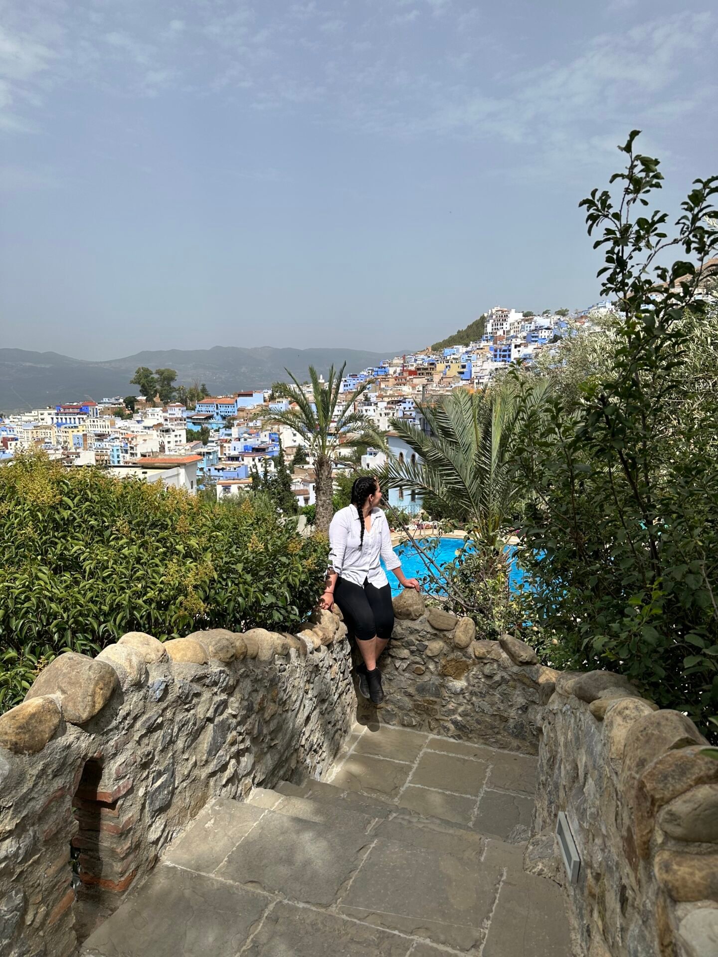 Seated comfortably, a figure enjoys a moment of tranquility with the captivating backdrop of Chefchaouen, Morocco. The city's distinctive blue-washed buildings create a striking contrast against the rugged landscape, while the distant mountains add depth to the scene. A harmonious blend of relaxation and vibrant culture, perfectly framed in this picturesque moment.