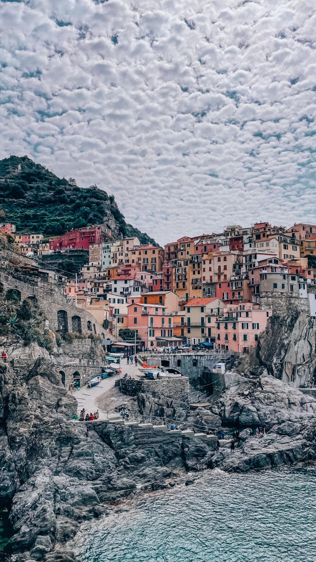 Under the vibrant summer sun, the iconic colorful houses of Cinque Terre cling to the rugged cliffside, each hue a testament to the coastal charm of the Italian Riviera. Above, fluffy white clouds form a picturesque dance across the azure sky, casting playful shadows on the picturesque scene. A harmonious blend of nature's beauty and human ingenuity, capturing the essence of a perfect summer day in Cinque Terre.