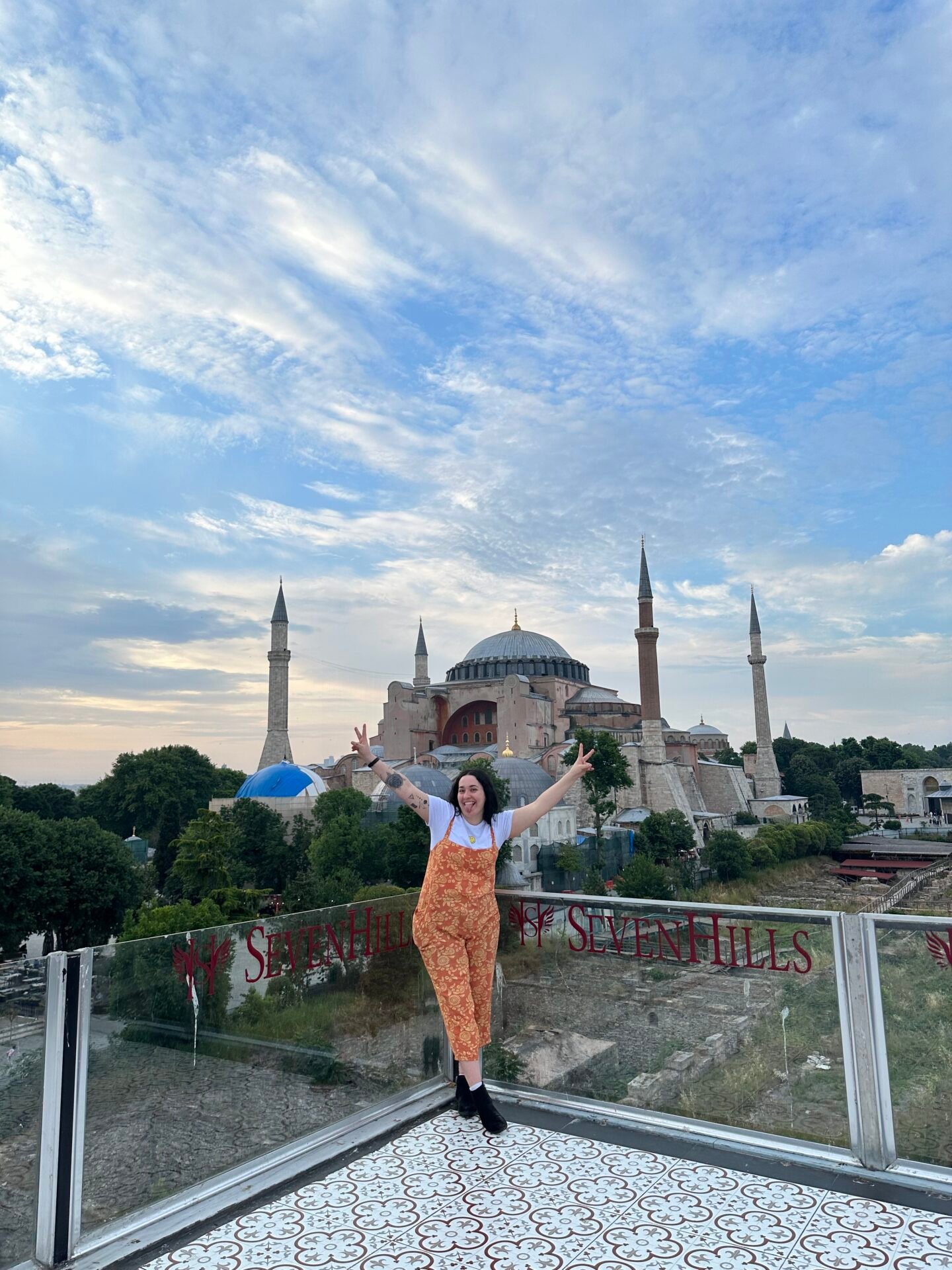 Vibrant bazaars and minarets adorn the bustling streets of Istanbul, Turkey. In the foreground, a girl stands on a balcony at Seven Hills resort and while in the distance, the iconic silhouette of the Blue Mosque graces the skyline against a backdrop of cloudy blue skies. A harmonious blend of ancient history and modern energy, this photo captures the essence of traveling in Turkey.