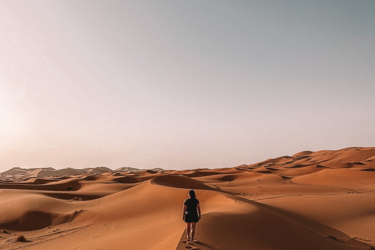 Standing on the red sands of the Sahara Desert, a figure gazes out over the vast expanse, where towering dunes stretch towards the horizon. The warm hues of the sand create a mesmerizing contrast with the clear blue sky, encapsulating the allure of this iconic desert landscape.